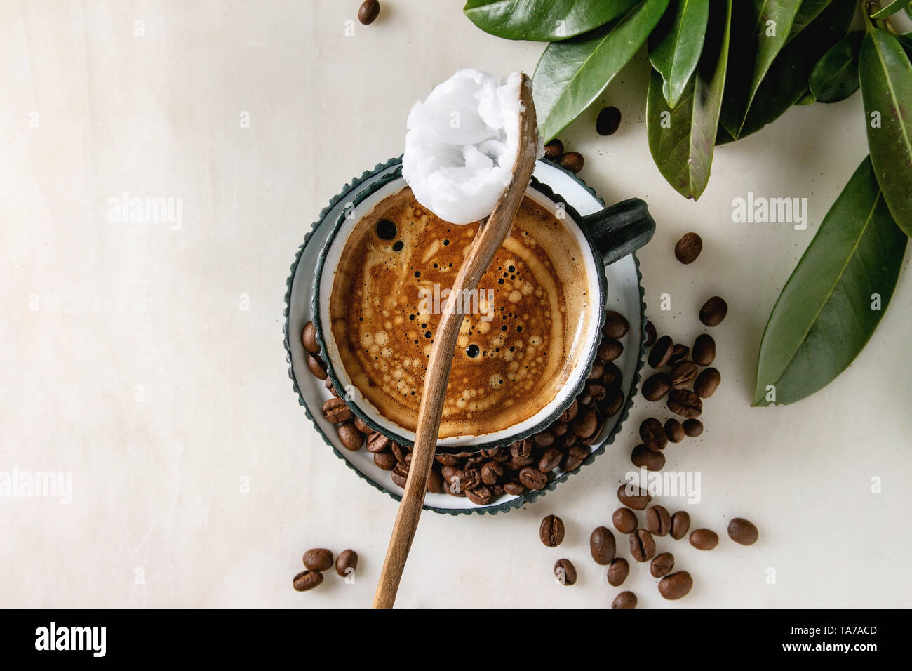 Bulletproof coffee. Keto diet coffee in blue ceramic cup with organic coconut cold press oil in wooden spoon with beans and green branch over white ma Stock Photo