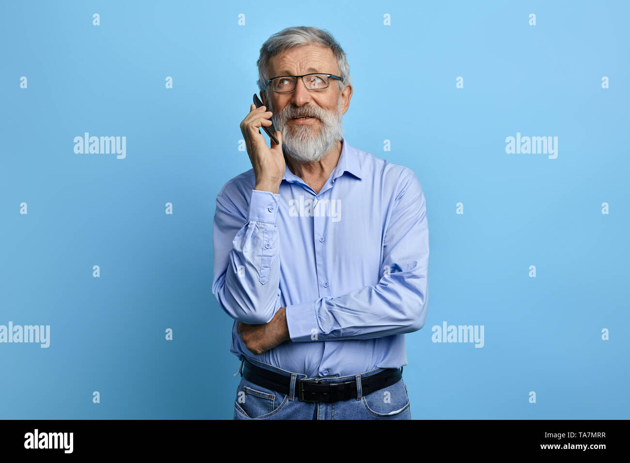handsome senior man looking up, talking on mobile phone isolated against sky blue background. old businessman having chat, business conversation witj  Stock Photo
