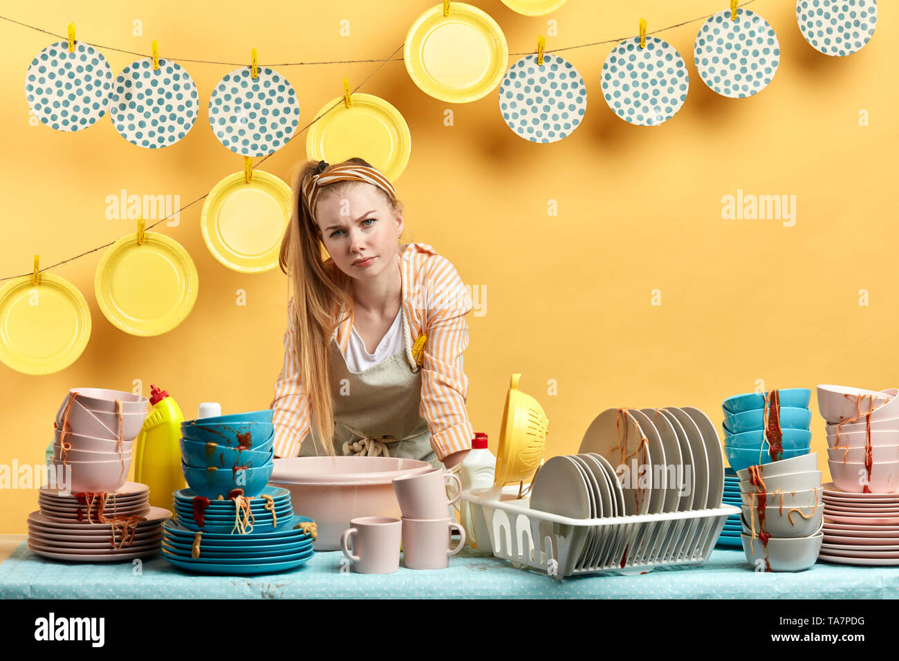 displeased unhappy housewife standing behind the kitchen table full of dirty tablewear. tiredness, laziness concept Stock Photo