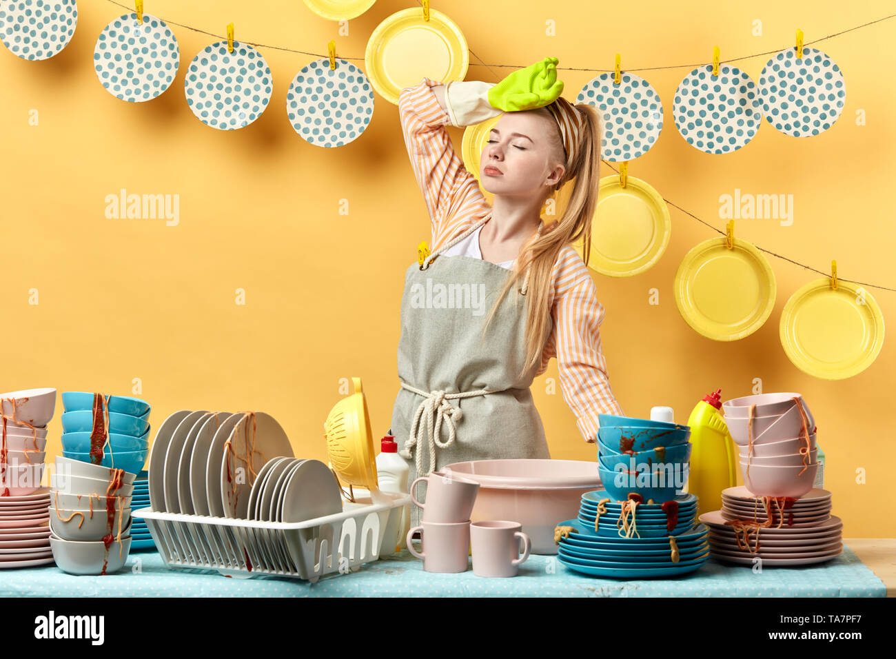 housewife holding her head, having headache, girl having a break, having a rest while washing the dishes in the kitchen with yellow wall, housewife ov Stock Photo