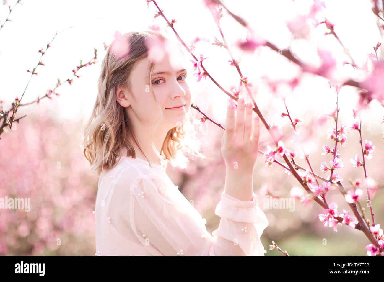 Smiling teen girl 16-17 year old posing in peach flowers outdoors. Looking at camera. Summer time. Stock Photo