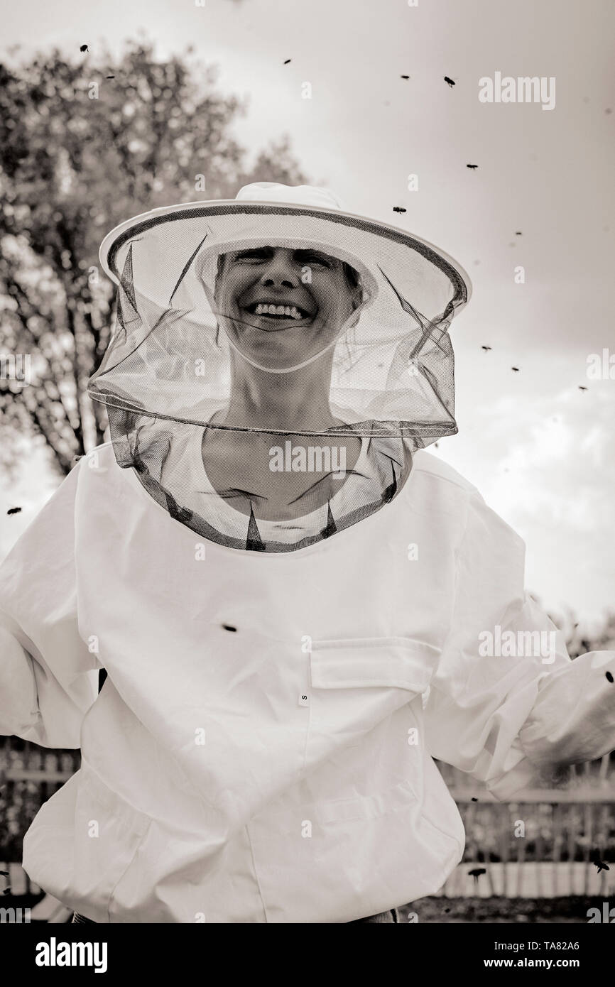 A smiling Beekeeper woman / apiarist portrait wearing a white smock with a hat and veil while working with her bees - beekeeping / apiculture. B/W Stock Photo