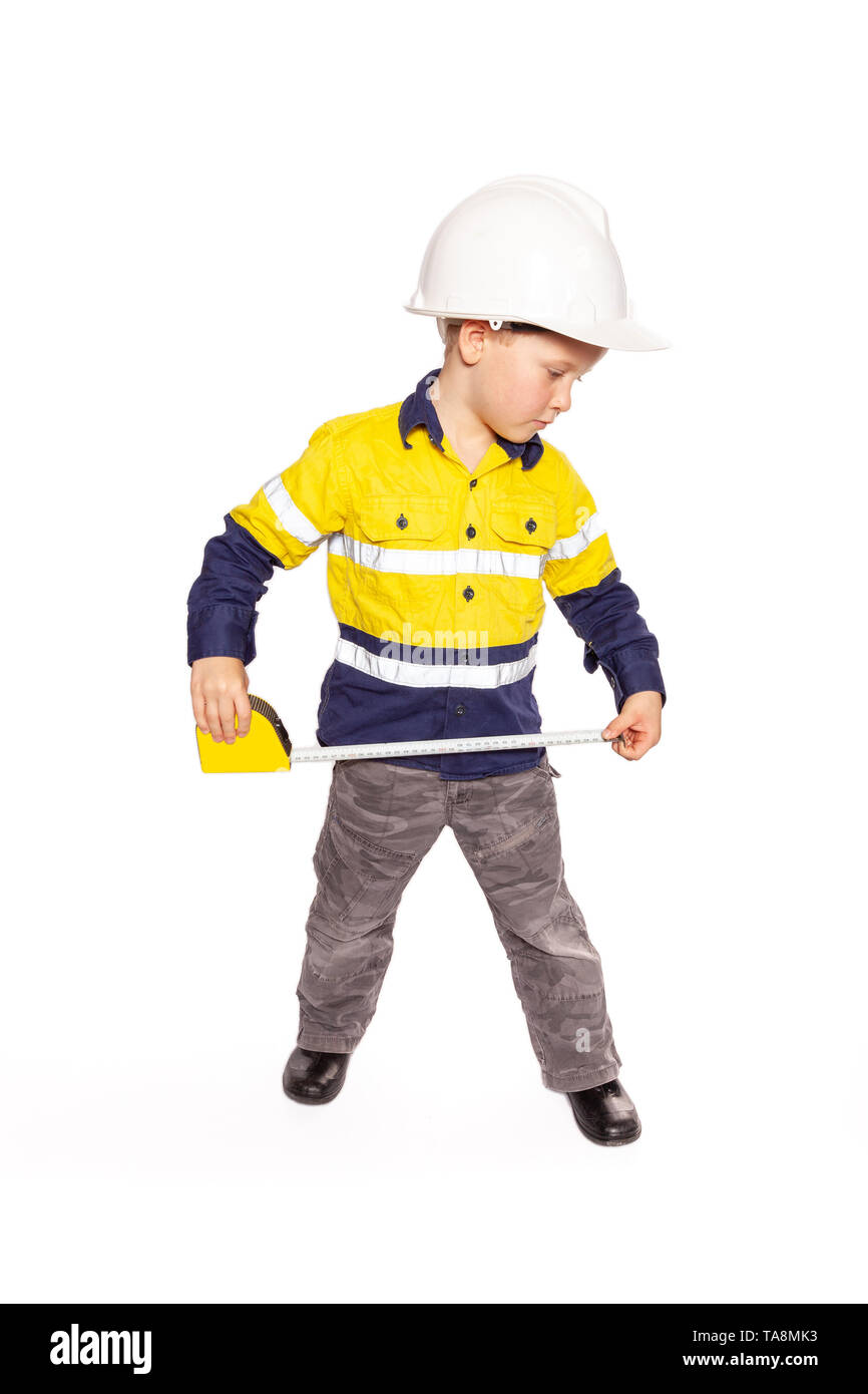 Young blond caucasian boy measuring something role playing as a construction worker in a yellow and blue hi-viz shirt, boots, white hard hat, and tape Stock Photo