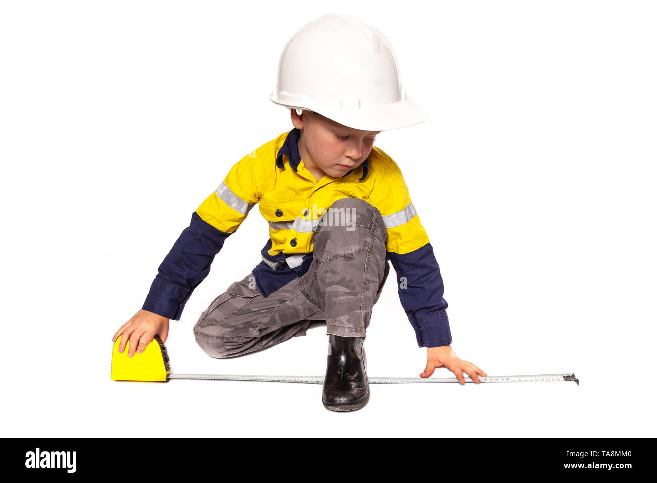 Young blond caucasian boy measuring something role playing as a construction worker in a yellow and blue hi-viz shirt, boots, white hard hat, and tape Stock Photo