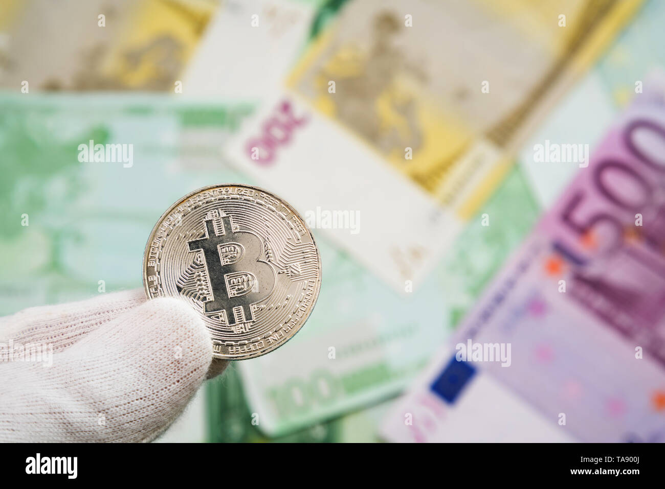 Man in white cloves holding Bitcoin coin between fingers with Euro bank notes in the background. Digital currency, block chain market Stock Photo