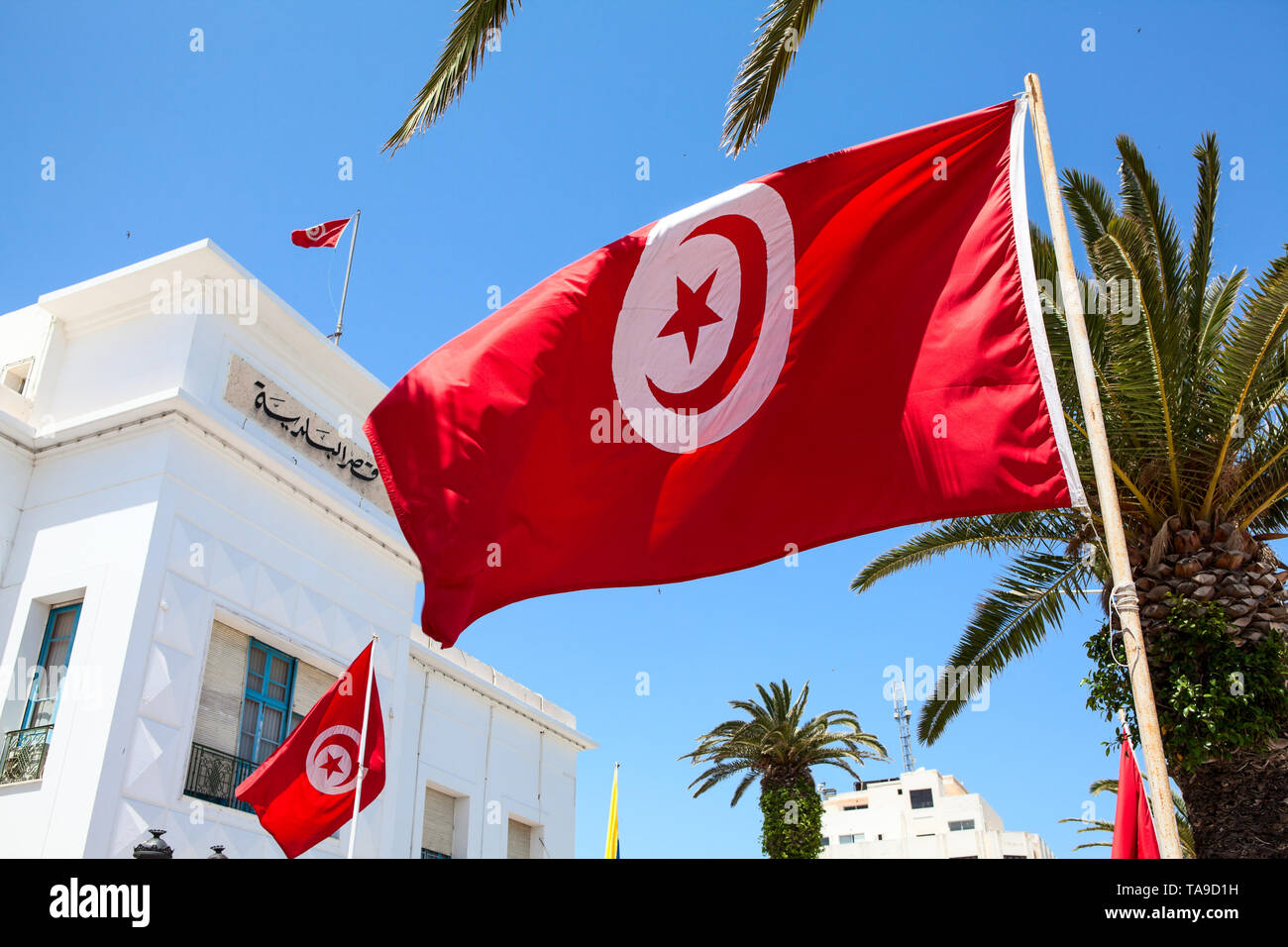 Tunisian national red flag with emblem crescent and star, against blue sky, urban background. The Tunisia, Africa Stock Photo