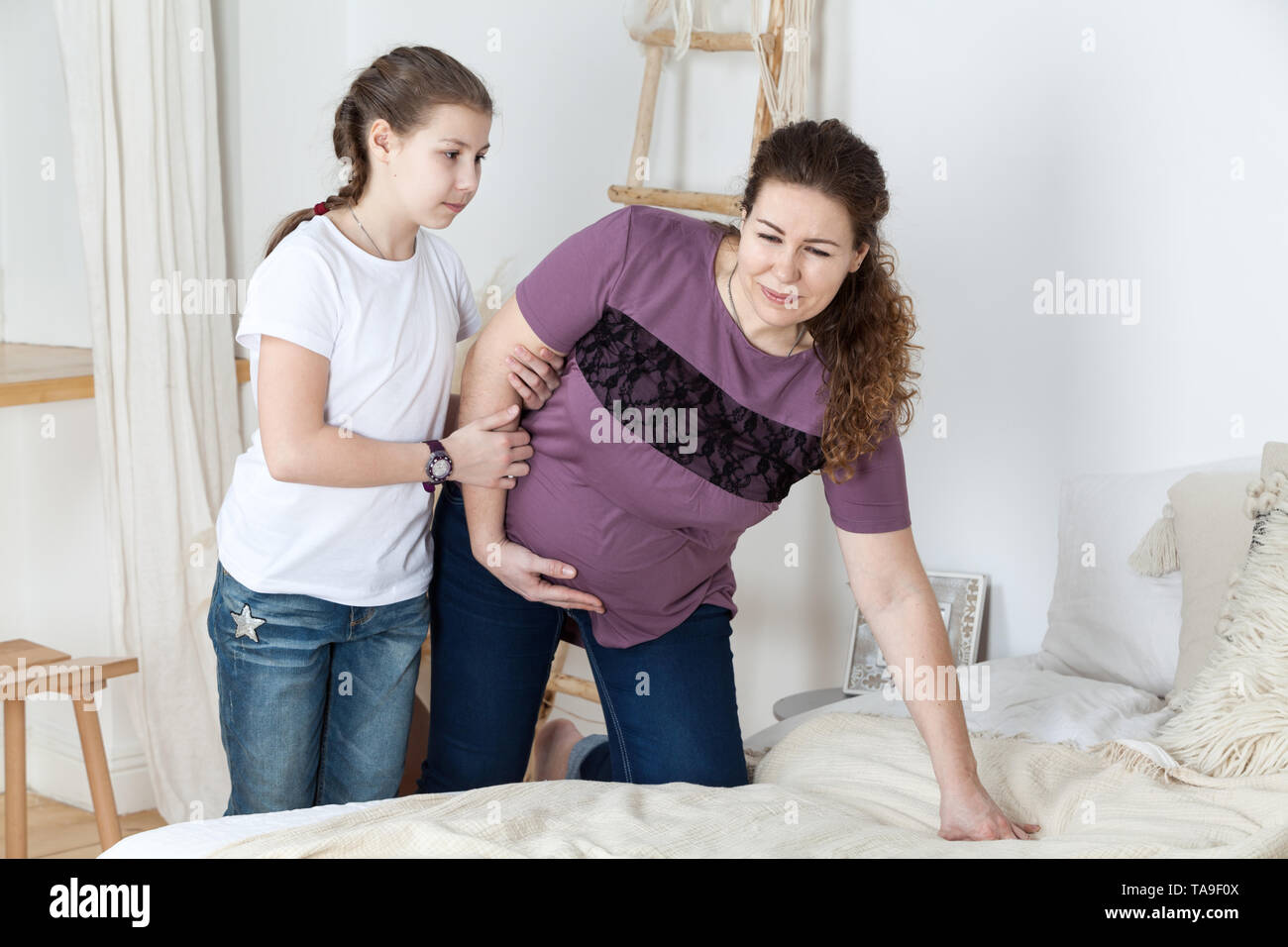 Teen daughter helping her pregnant mother to lie down on bed, stomach