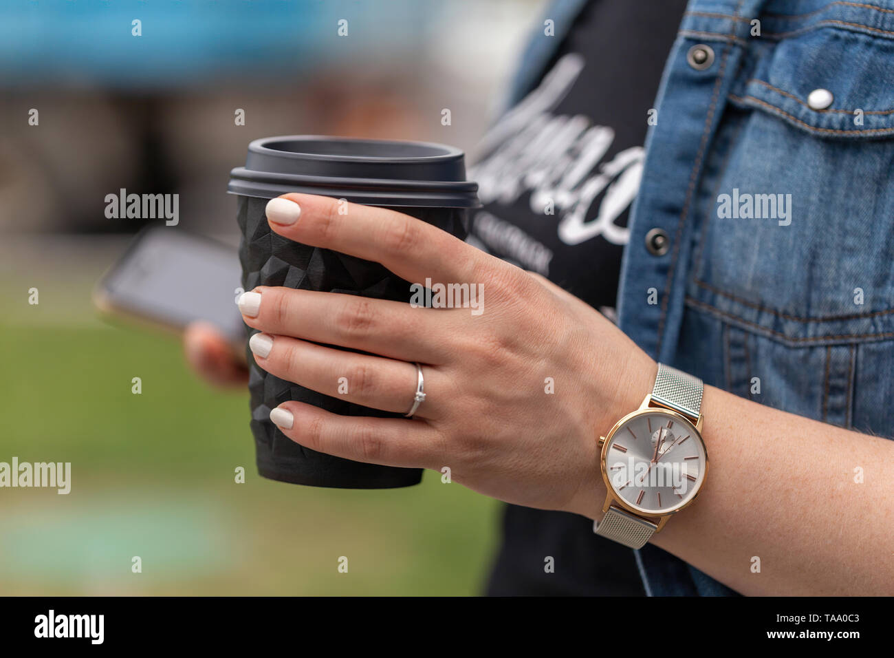Close up of women's hands holding smartphone and a cup of coffee. A woman is dressed in a blue denim jacket, a black T-shirt and a watch on her arm Stock Photo