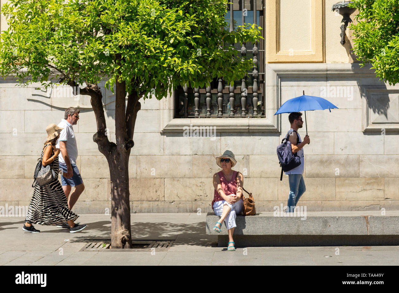 Woman sitting on a bench in Seville, Andalusia region, Spain. A man with a blue sun umbrella walking by. Stock Photo