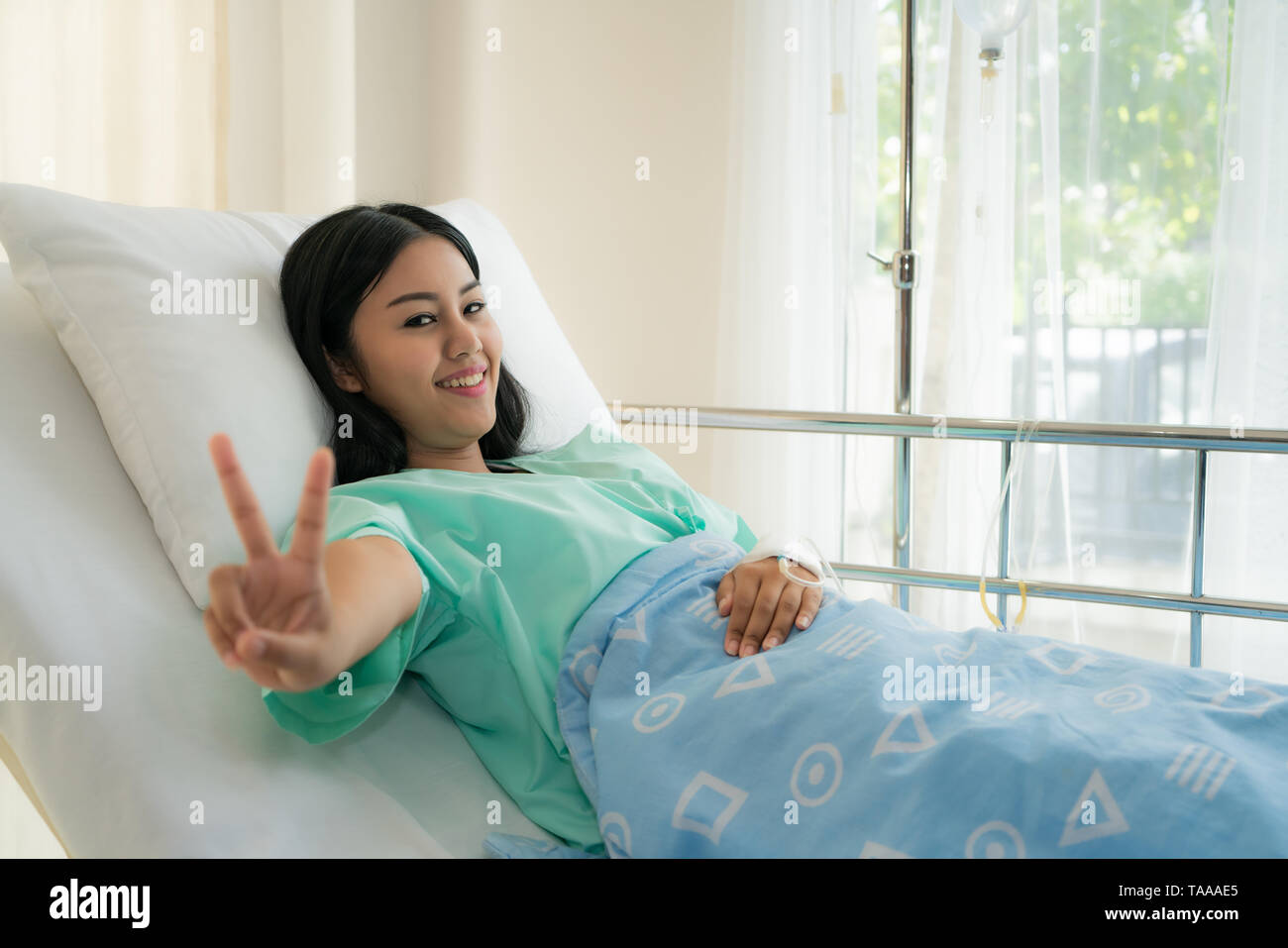 Young Asian patient woman lying at hospital bed with saline drip showing victory sign for cheerful. Drop of saline solution to help patient. Stock Photo