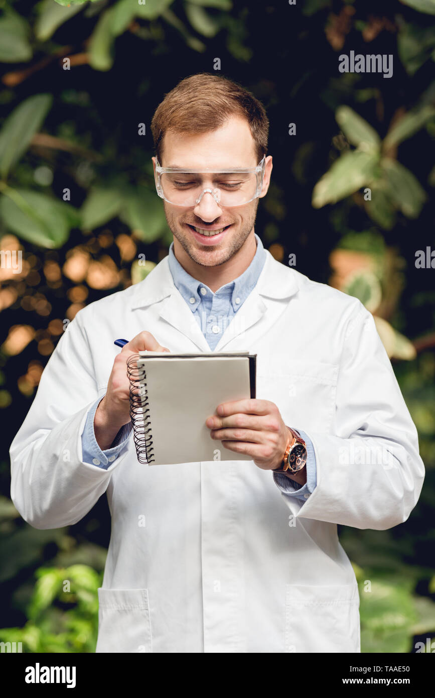 smiling scientist in white coat and goggles making notes in green orangery Stock Photo