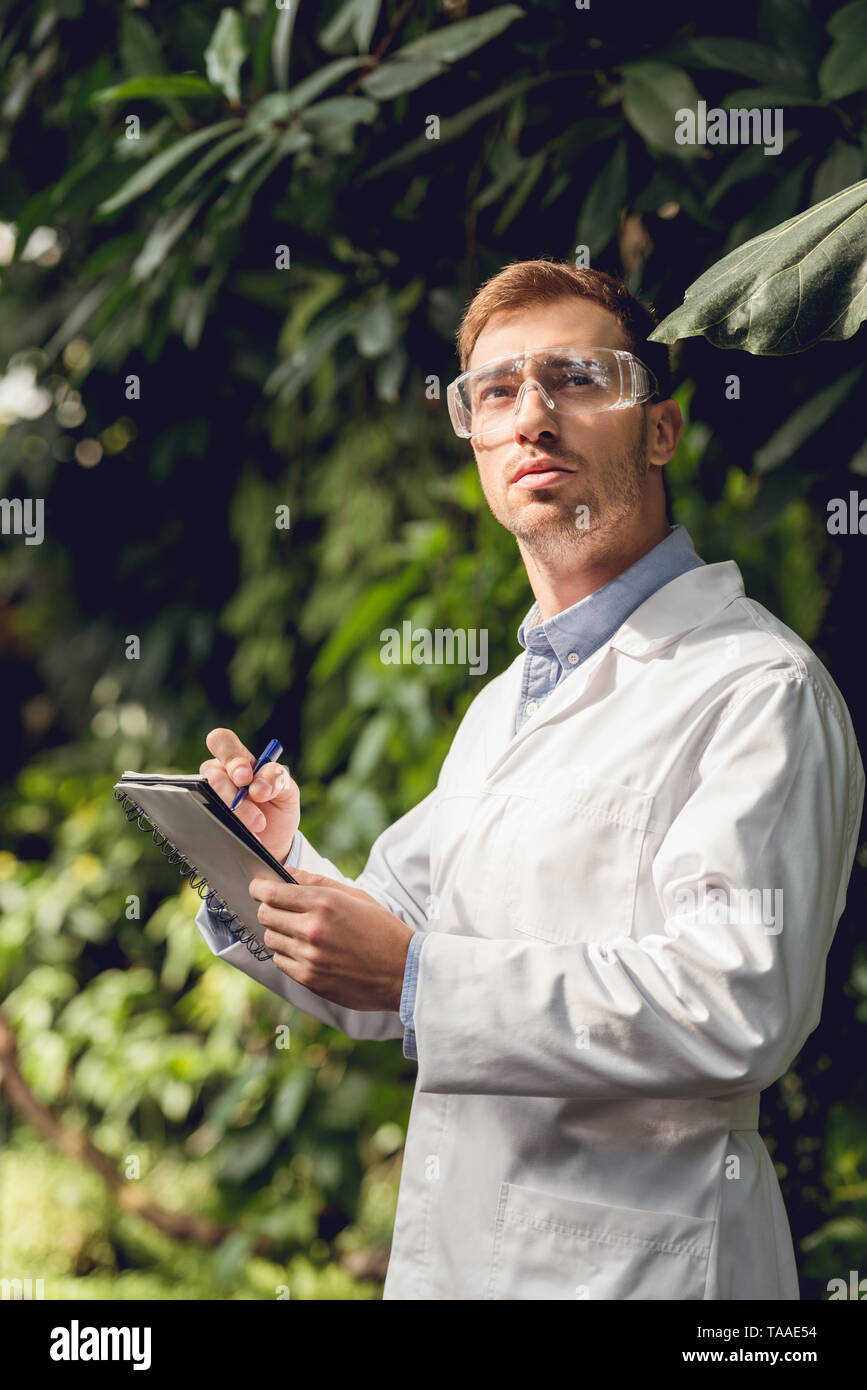 scientist in white coat and goggles making notes in green orangery Stock Photo