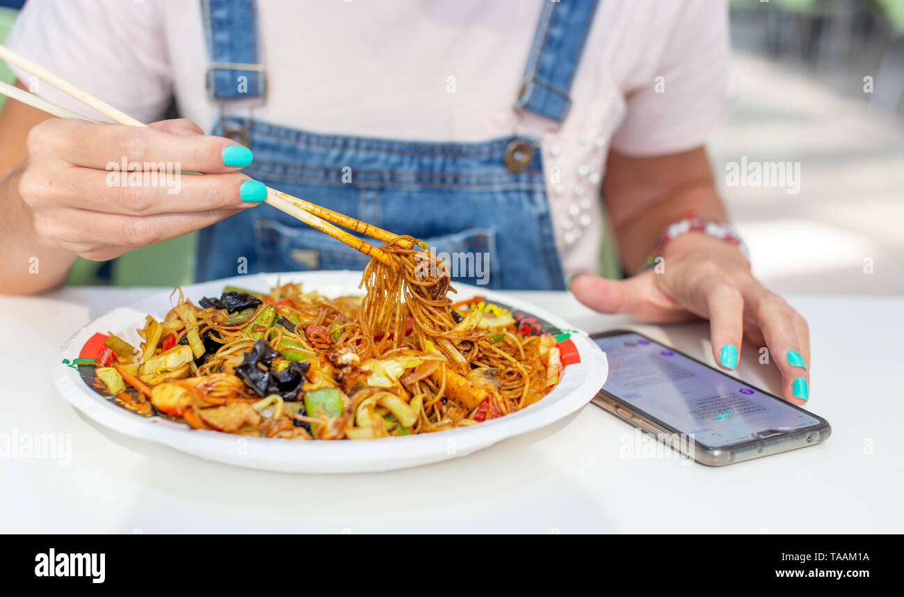Woman eating chinese noodle with chicken and messaging on smartphone closeup Stock Photo