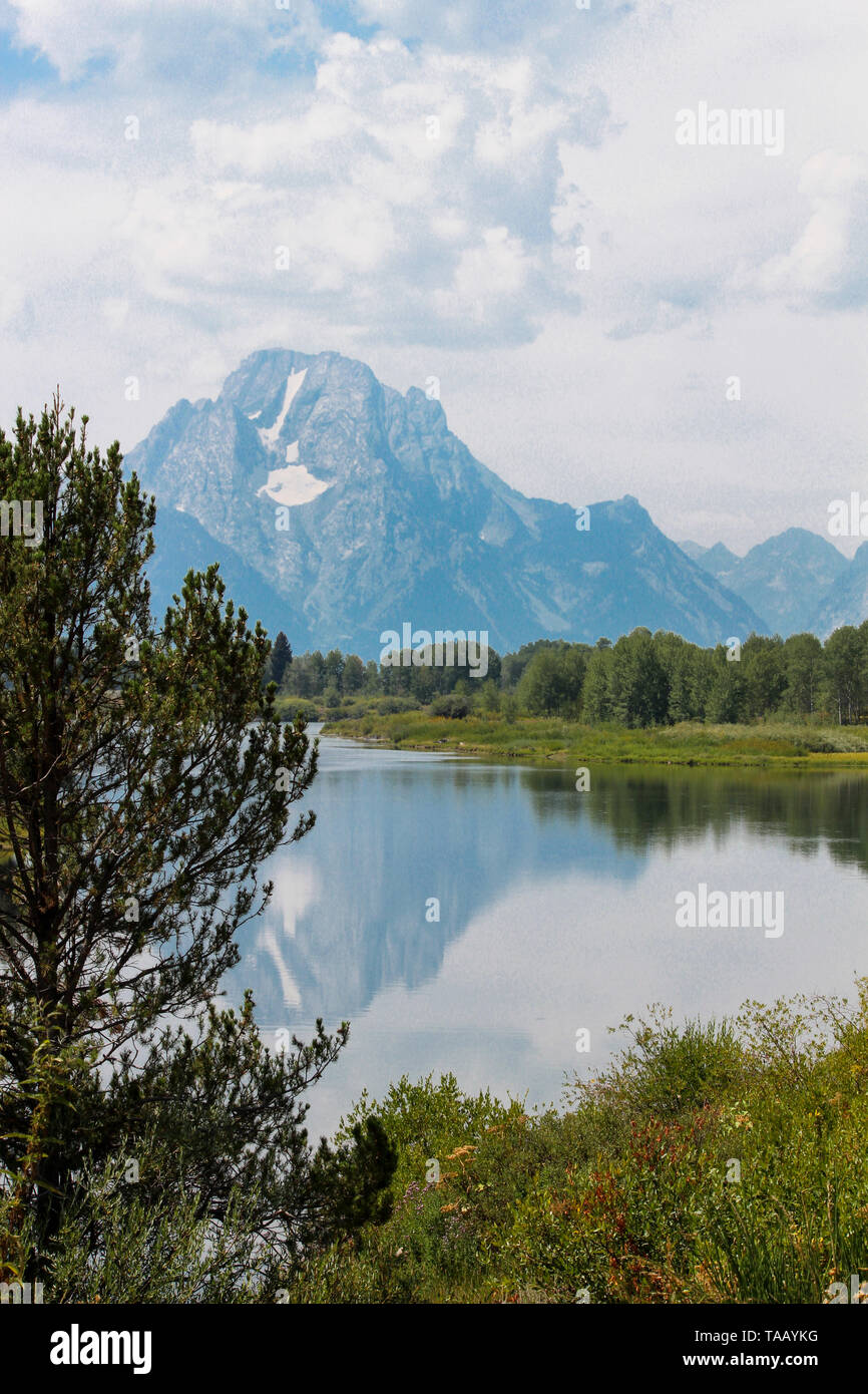 Scenic landscape near Grand Tetons and Yellowstone National Parks, Wyoming.  Mountains with clouds in blue sky, with lake and tree in foreground. Stock Photo