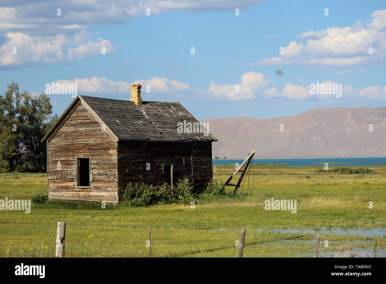 Rustic brown cabin in rural farm landscape near lake with mountains in distance, under a blue sky with clouds. Stock Photo