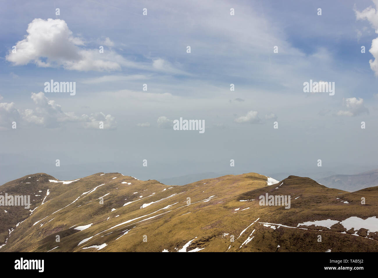 Blue sky with white clouds over sunlit highlands and grasslands of Old mountain with distant pointy peaks and remaining snow during early spring Stock Photo