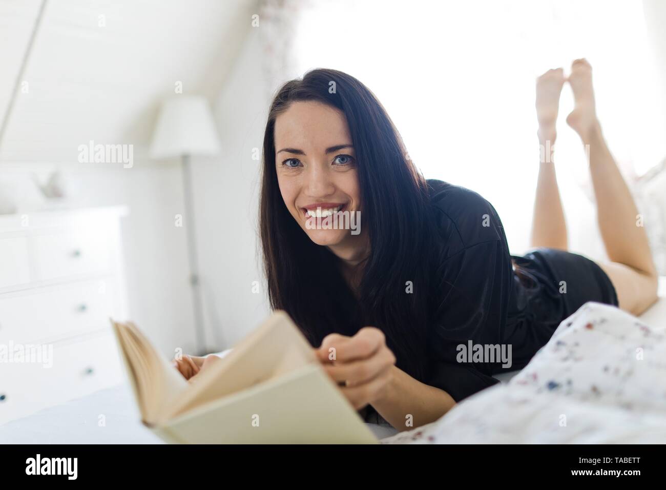 Black haired woman sleepwear laying on bed and reading book Stock Photo