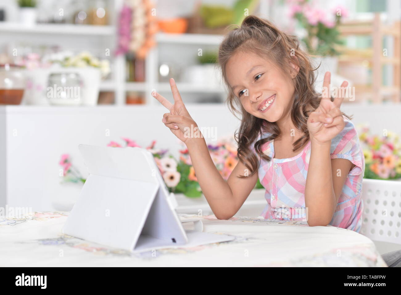 Portrait of cute little girl sitting at kitchen table in front of laptop and showing peace signs Stock Photo