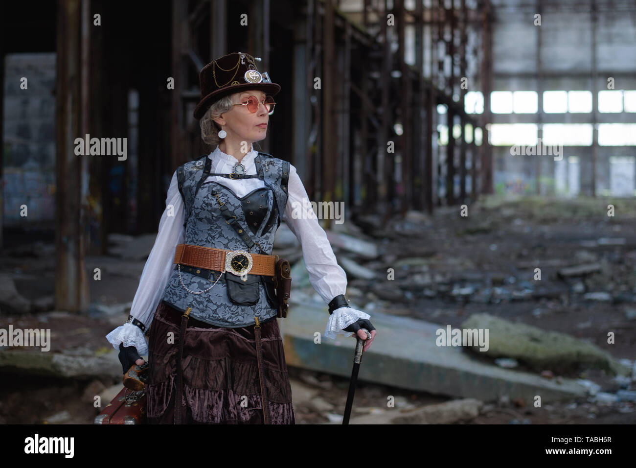 elderly lady in a steampunk costume at an abandoned factory with arms in hand. Stock Photo