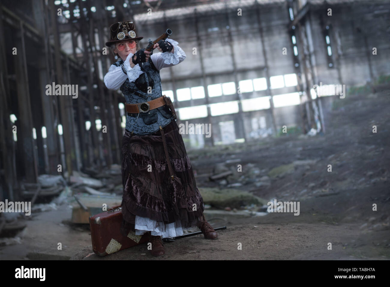 elderly lady in a steampunk costume at an abandoned factory with arms in hand. Stock Photo