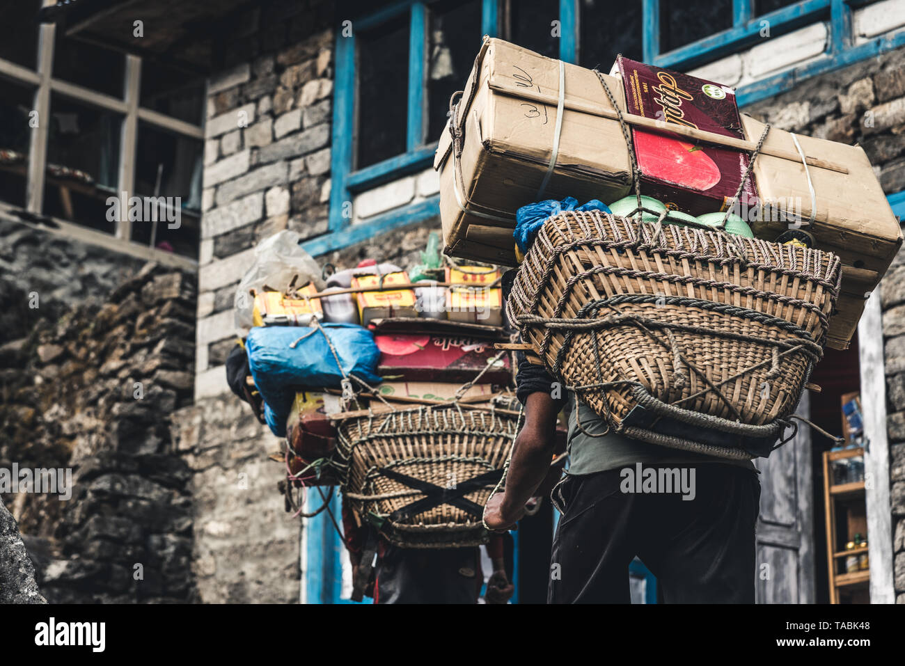 Low angle view of men walking through streets of small Nepalese village with large pack of supplies on his back. Stock Photo