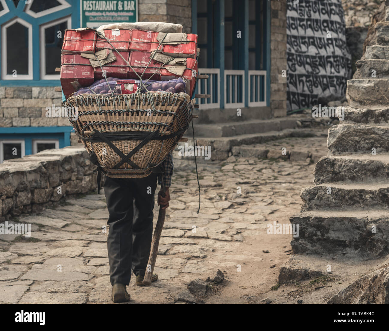 Man walking through streets of small Nepalese village with large pack of supplies on his back. Stock Photo