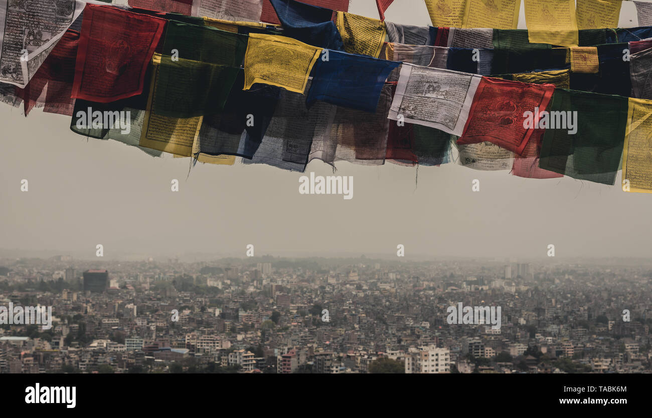 Colorful rectangular cultural prayer flags blowing in the wind against the backdrop of a Nepalese city far below. Stock Photo