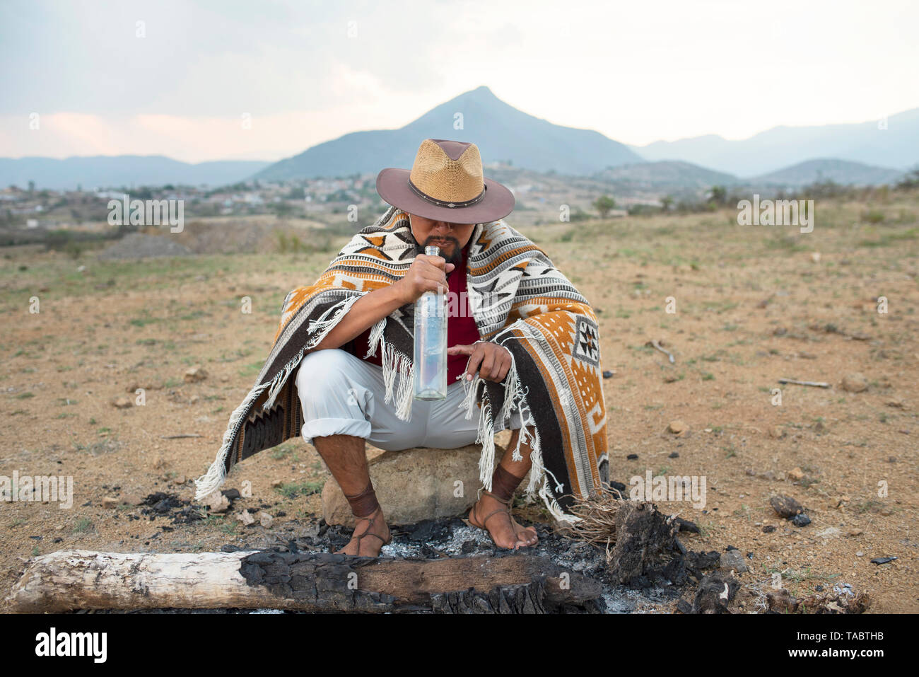 Zapotec (Mexican) man wearing a hat, Zapotec rug and ancient sandals drinking a bottle of Mezcal. Oaxaca State, Mexico Stock Photo