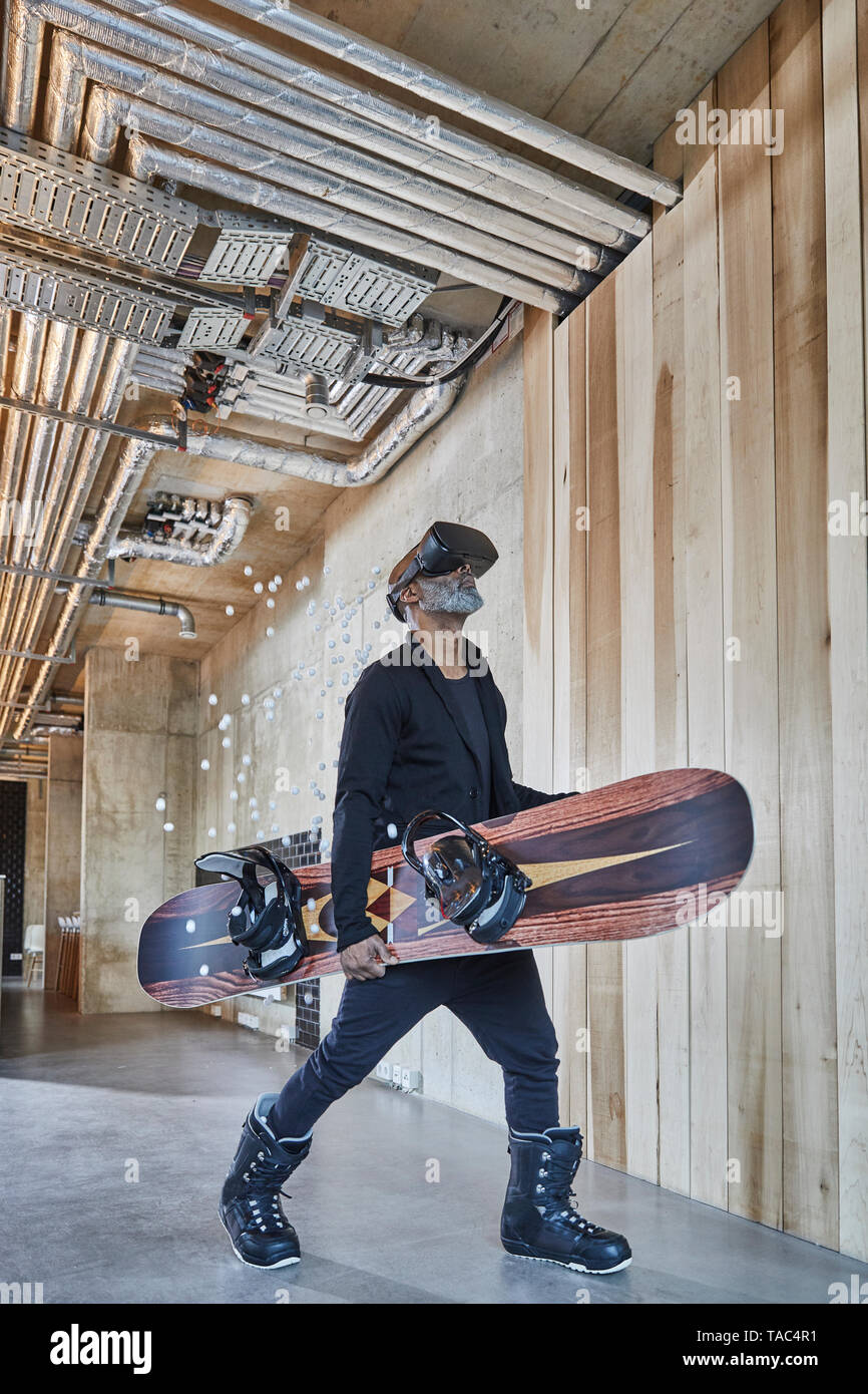 Mature businessman wearing VR glasses holding snowboard in modern office Stock Photo
