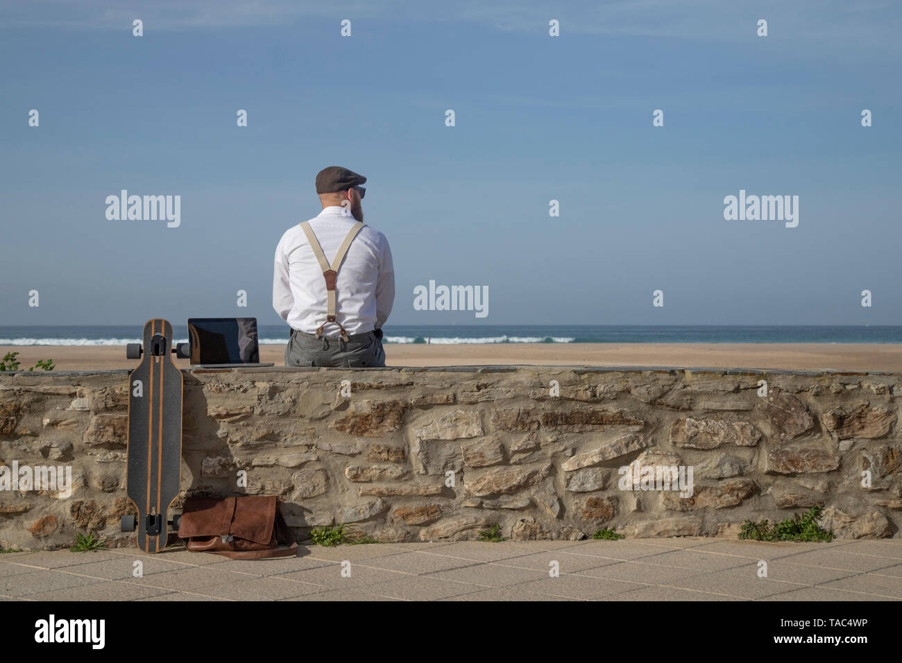 Back view of man with laptop and longboard sitting on a wall in front of beach and sea looking at distance Stock Photo