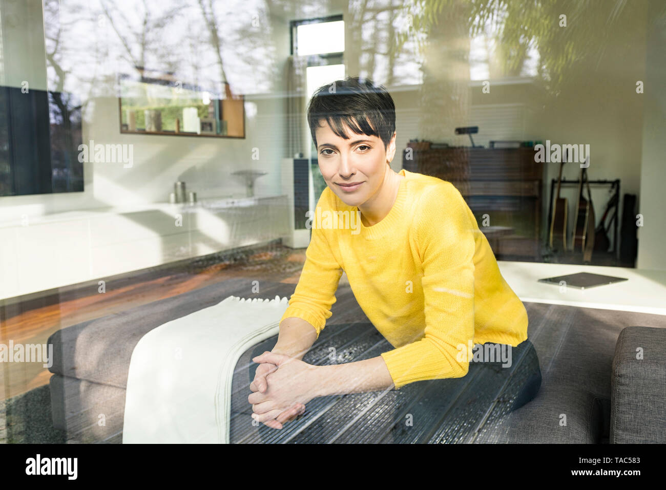 Portrait of content woman sitting behind windowpane on couch in the living room Stock Photo