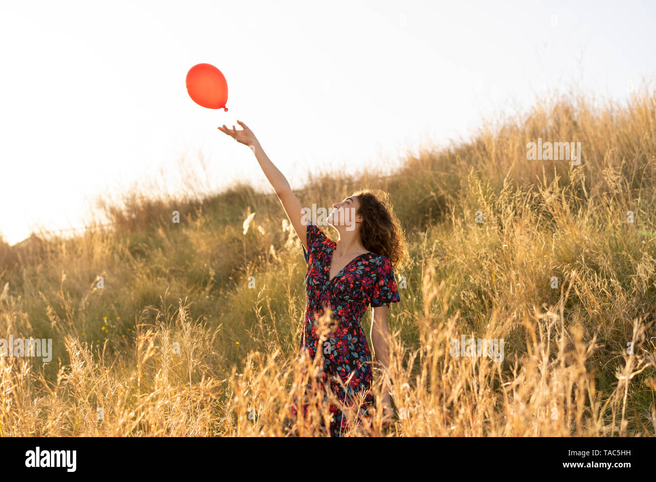 Happy young woman standing in summer meadow, letting go of a red balloon Stock Photo
