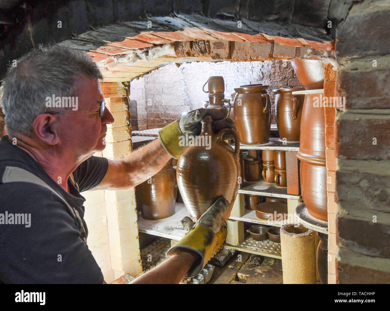 24 May 2019, Brandenburg, Groß Neuendorf: Manfred Dannegger, potter, crouches in front of his still hot oven and removes burnt jugs, cups and other vessels from the combustion chamber. Since 1997 the potter has been producing this clay glazed stoneware in the tradition of Bunzlau brownware. The dishes are fired in the oven at about 1300 degrees Celsius for about 20 hours. The oven must then cool for four days. This weekend (25.05./26.05.) the 12th pottery market will take place in Groß Neuendorf. Then 22 potters from near and far present themselves directly at the dike of the German-Polish bor Stock Photo