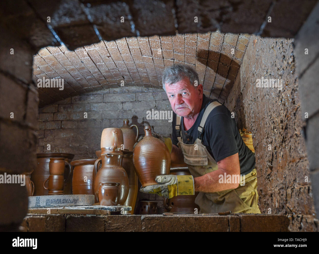 24 May 2019, Brandenburg, Groß Neuendorf: Manfred Dannegger, a potter, stands in his still hot oven and removes burnt jugs, cups and other vessels from the combustion chamber. Since 1997 the potter has been producing this clay glazed stoneware in the tradition of Bunzlau brownware. The dishes are fired in the oven at about 1300 degrees Celsius for about 20 hours. The oven must then cool for four days. This weekend (25.05./26.05.) the 12th pottery market will take place in Groß Neuendorf. Then 22 potters from near and far present themselves directly at the dike of the German-Polish border river Stock Photo