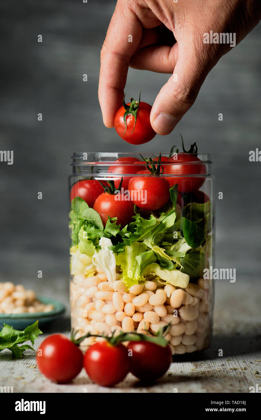 closeup of a young caucasian man preparing a jar salad, with cherry tomatoes, lettuce and cooked white beans, on a rustic wooden table against a gray  Stock Photo
