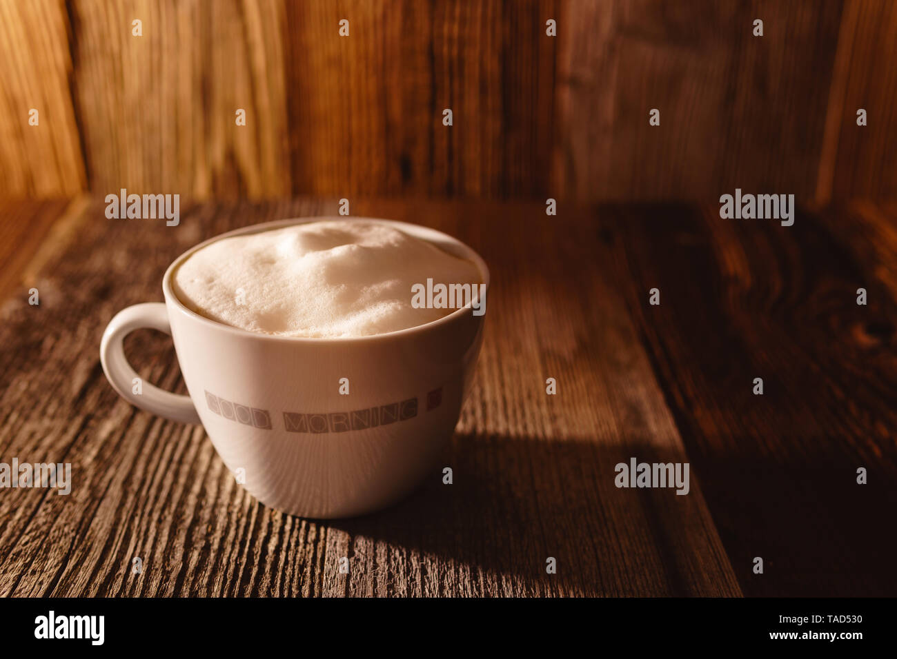 Freshly brewed coffee topped with vegan soy milk foam in a white cup with rustic brownish wooden background in beautiful moody morning sunlight Stock Photo