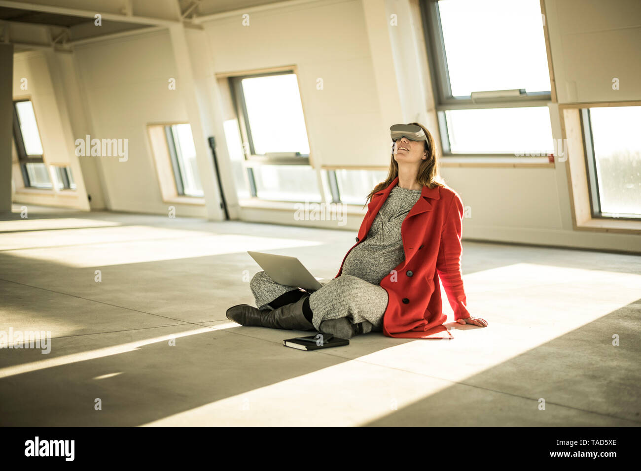 Pregnant busnesswoman sitting on floor of new office rooms, using VR goggles and laptop Stock Photo