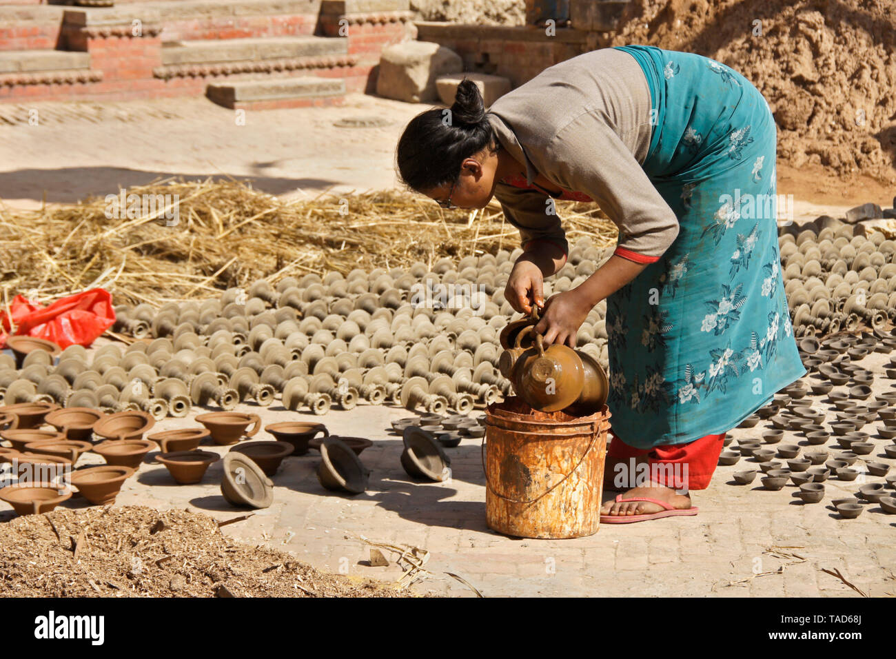 Woman dipping pottery into color as other clay products sun-dry in Kumale Tol (Potters' Square, Pottery Square), Bhaktapur, Kathmandu Valley, Nepal Stock Photo