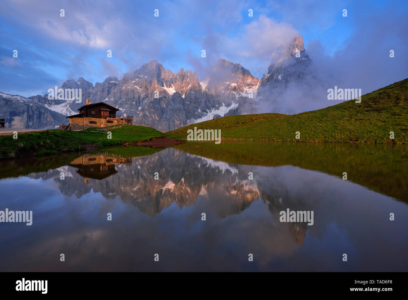 Italy, Trentino, Dolomites, Passo Rolle, Pale di San Martino range, Cimon della Pala with Baita Segantini reflecting in small lake in the evening Stock Photo