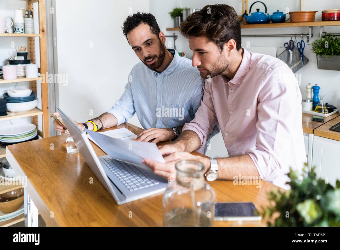 Two young men working together in kitchen, using laptop, discussing documents Stock Photo