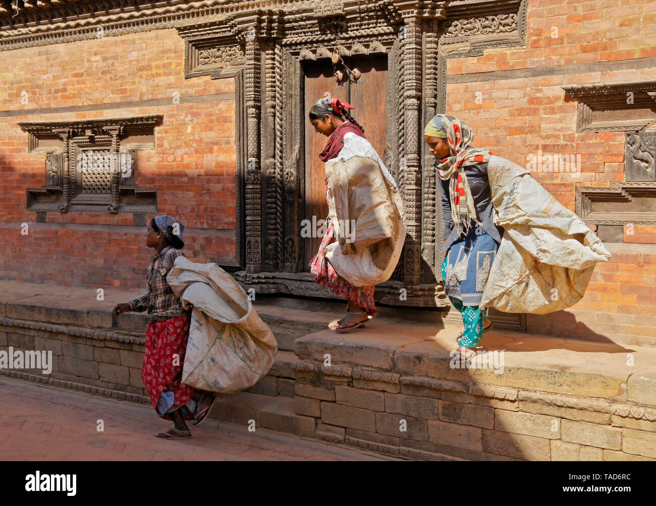 Female workers carrying large bags past carved wood door of old brick building in Durbar Square, Bhaktapur, Kathmandu Valley, Nepal Stock Photo