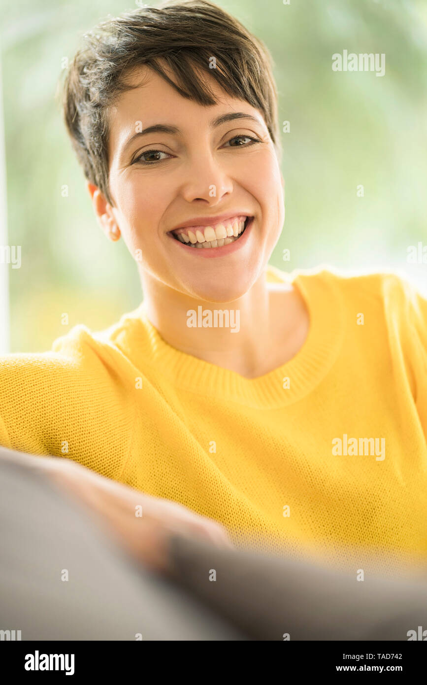 Portrait of happy woman with short brown hair wearing yellow pullover sitting on the couch at home Stock Photo