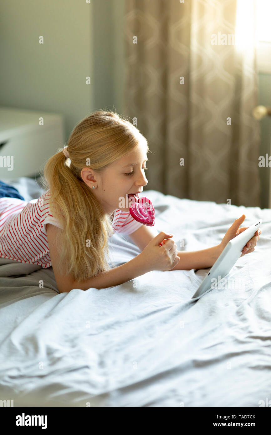 Blond girl lying on bed licking lollipop while using digital tablet Stock Photo