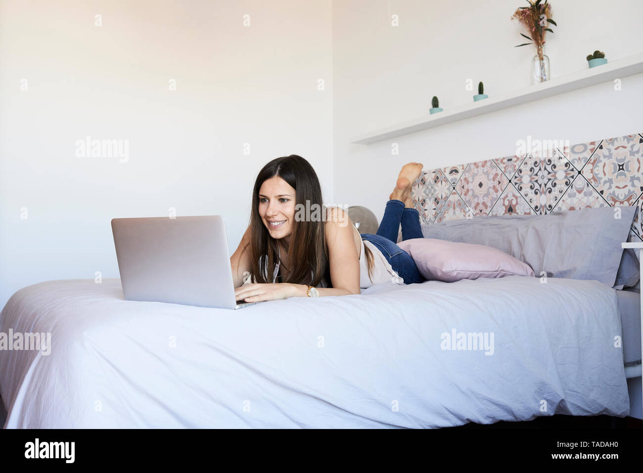 Portrait of smiling young woman lying on bed using laptop Stock Photo ...