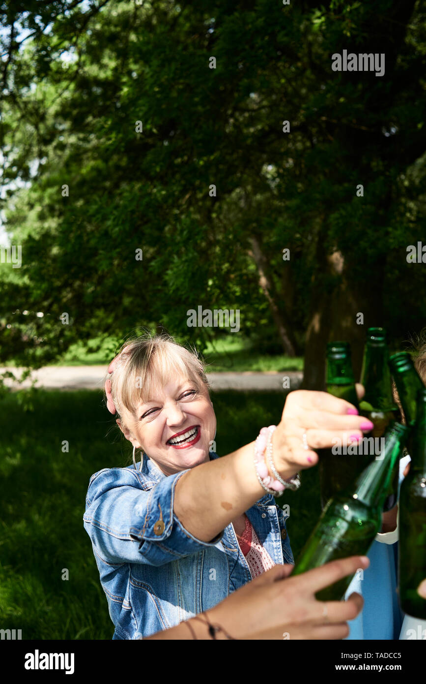 Portrait of happy woman clinking beer bottle with friends in park Stock Photo