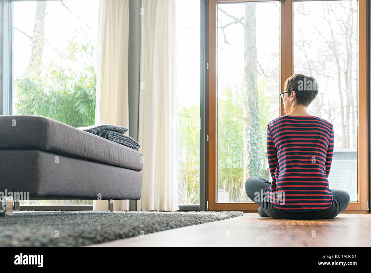 Back view of woman sitting on the floor of living room looking out of window Stock Photo