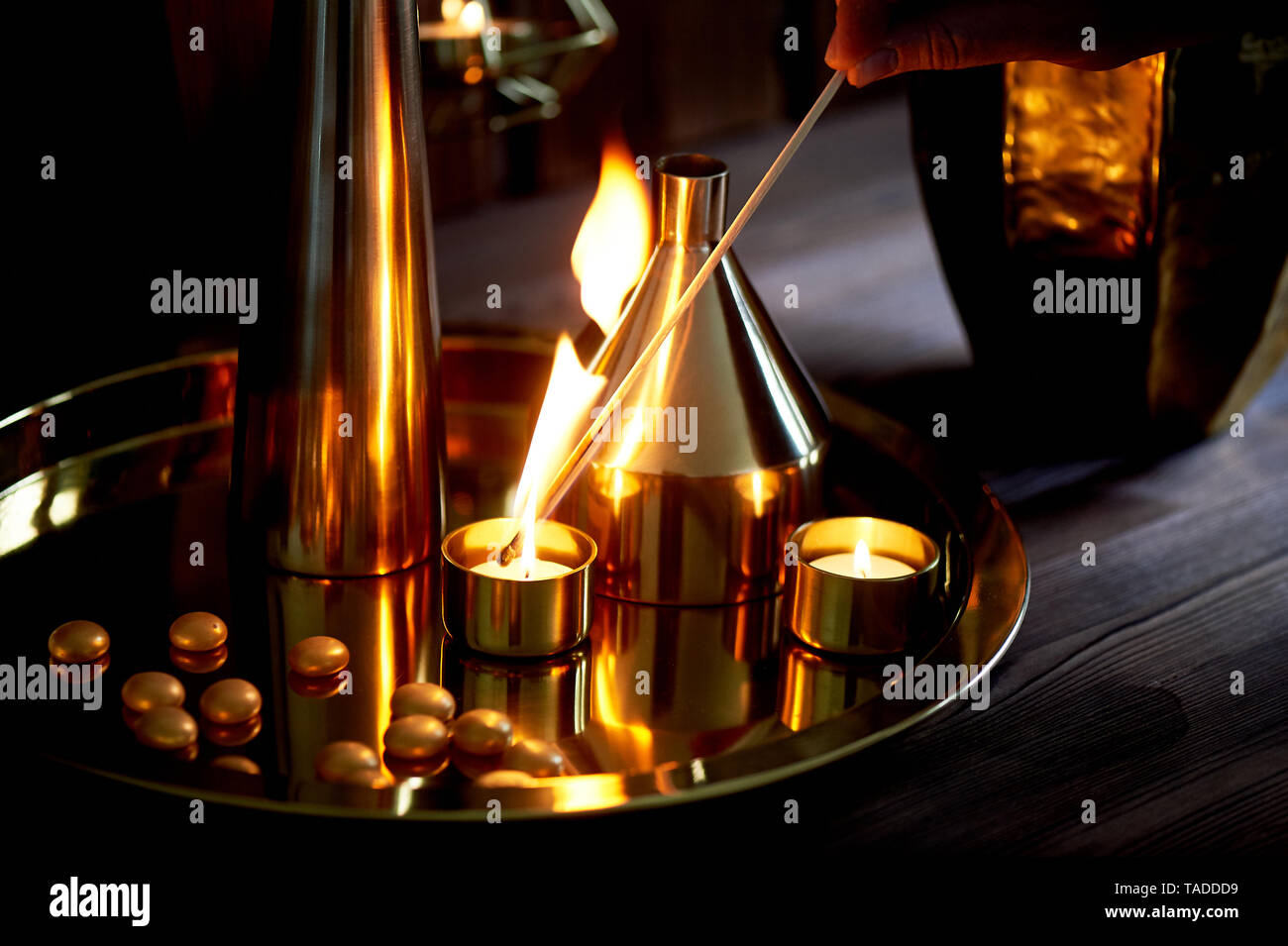 Hand lights a candle with a long match with a bright flame. The warm Golden gamma. Evening comfort. Many vases of different shapes of gold metal. Stock Photo