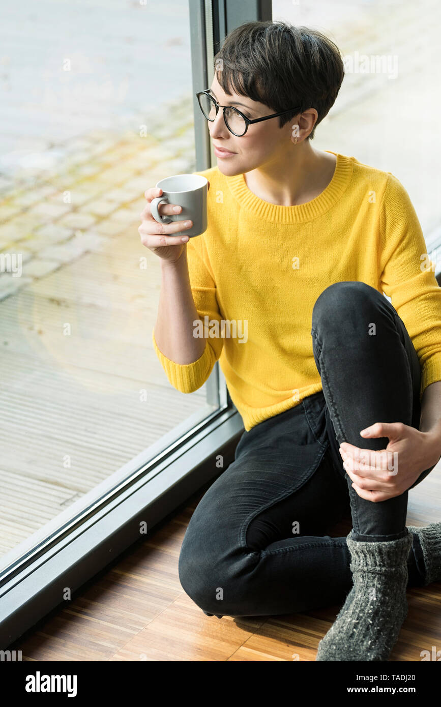 Woman with cup of coffee sitting on the floor at home looking out of window Stock Photo