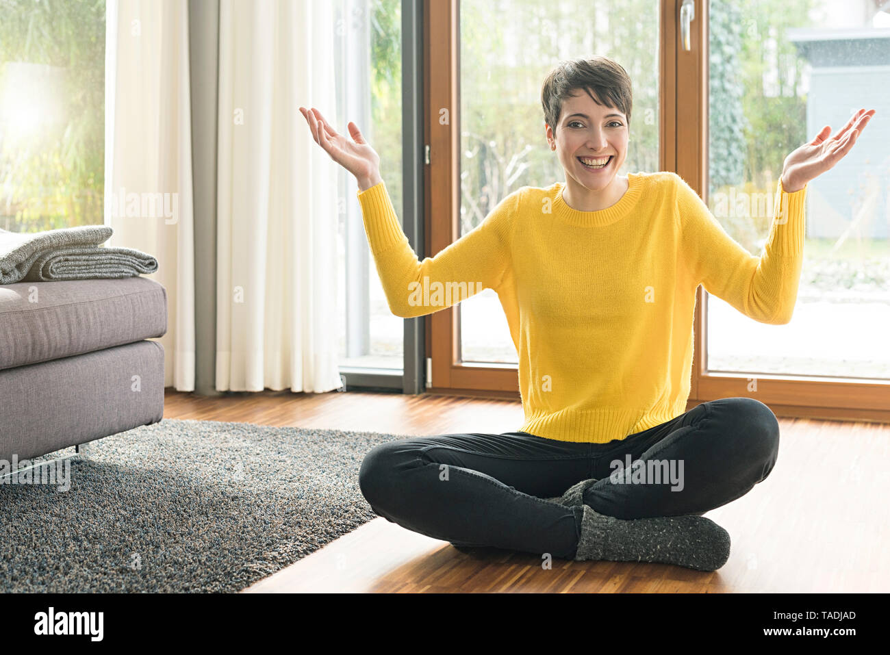 Portrait of happy woman sitting on the floor of living room with hands raised Stock Photo