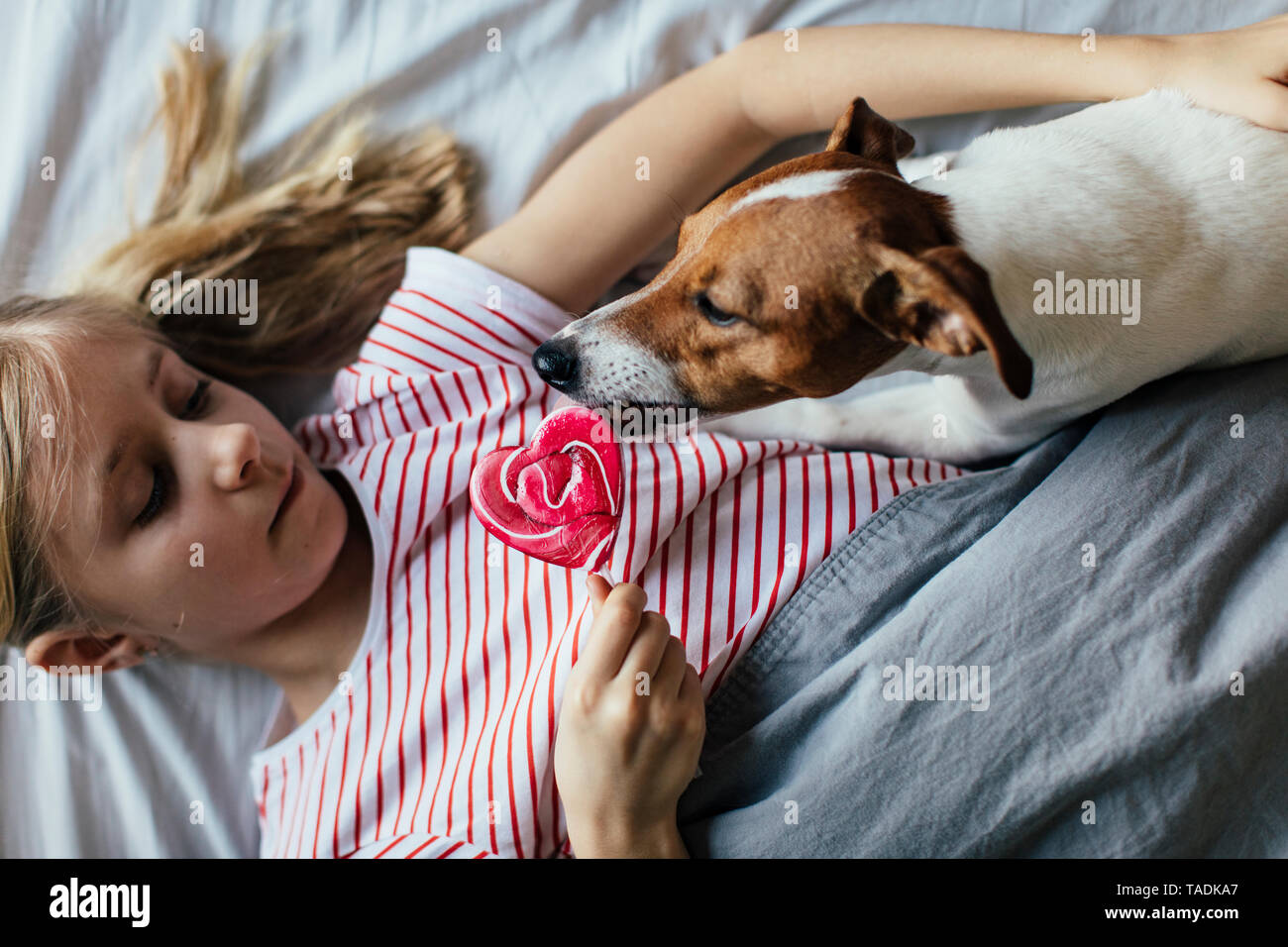 Girl lying on bed watching her dog licking lollipop Stock Photo
